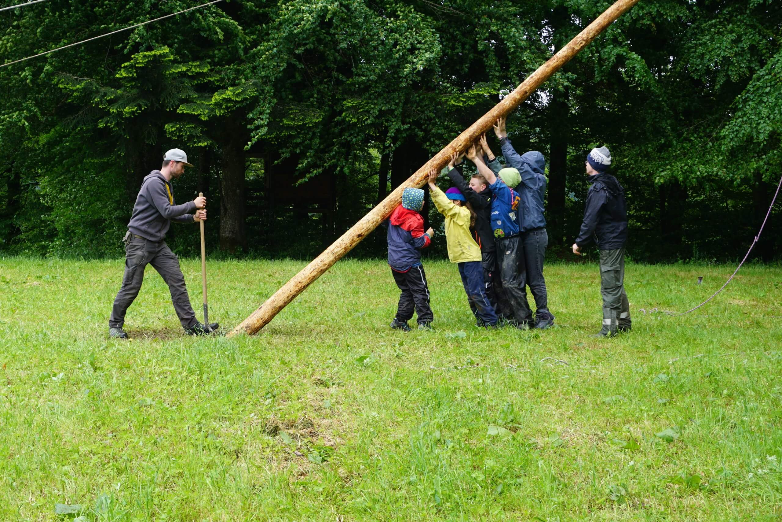 Group of children and adults in a grassy clearing work together to lift a tall, leaning log outdoors.