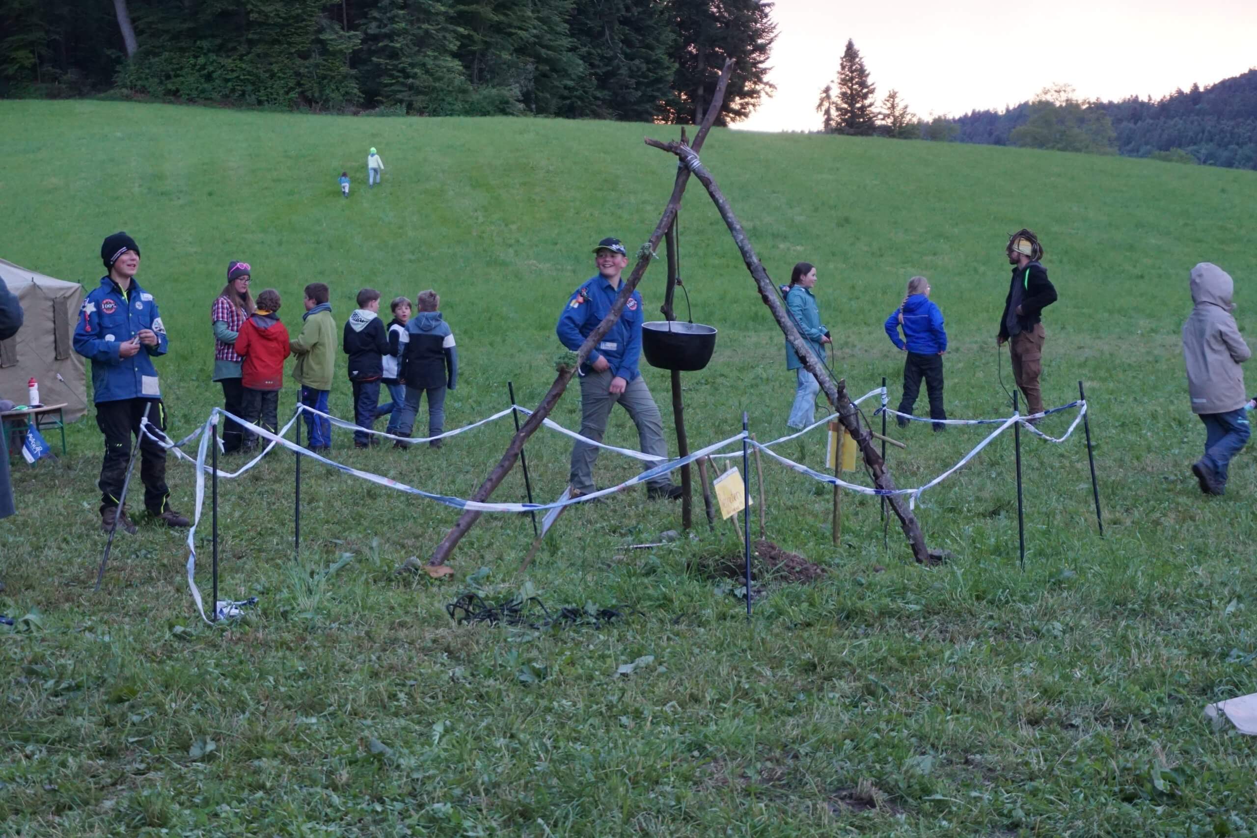 Group of kids and adults at a grassy field camp, with a tripod cooking setup and a tent in the background.