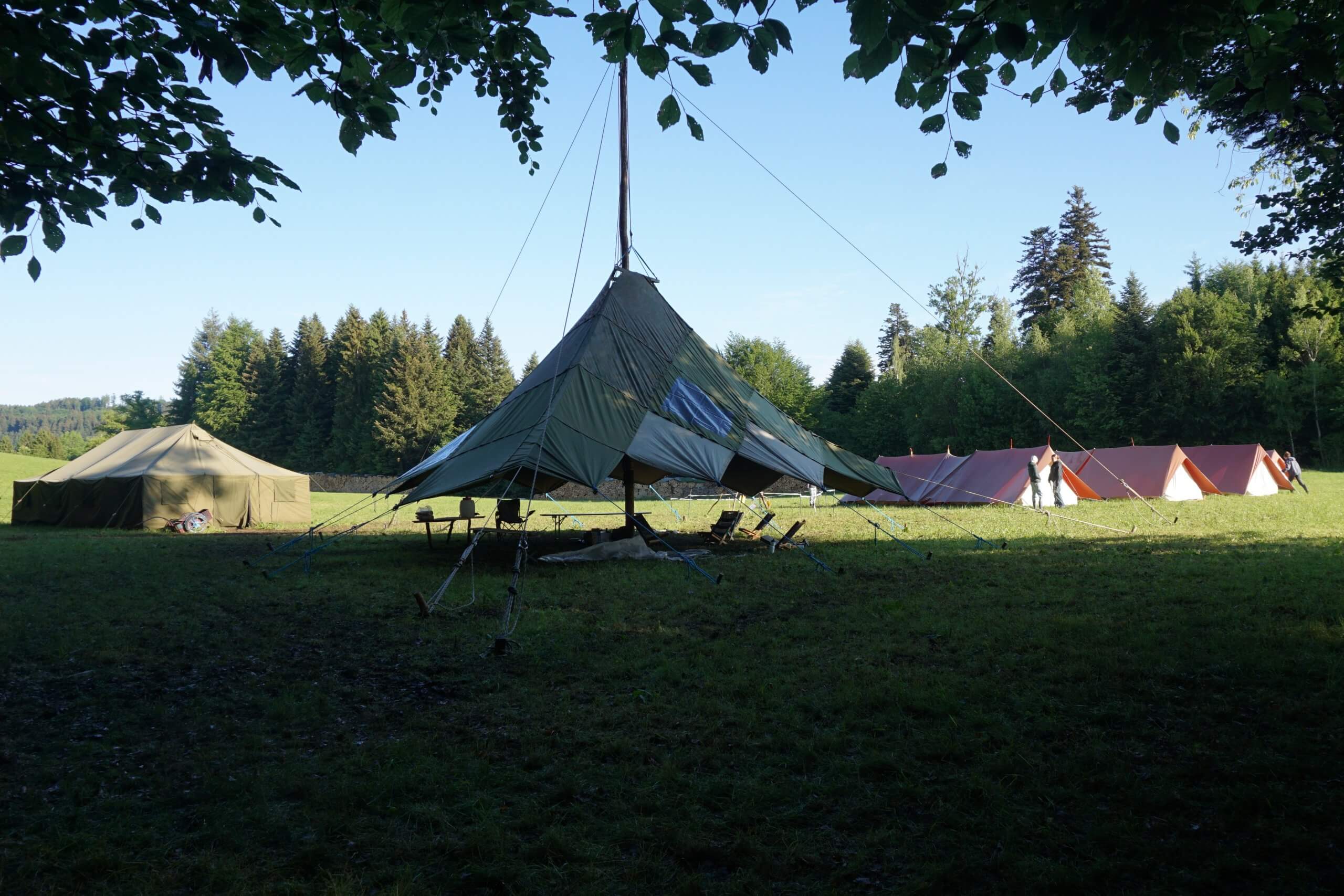 A large central ceremonial tent with tall poles and ropes set in a grassy campsite, with smaller canvas tents along the edge and trees in the background and a few people on the right.