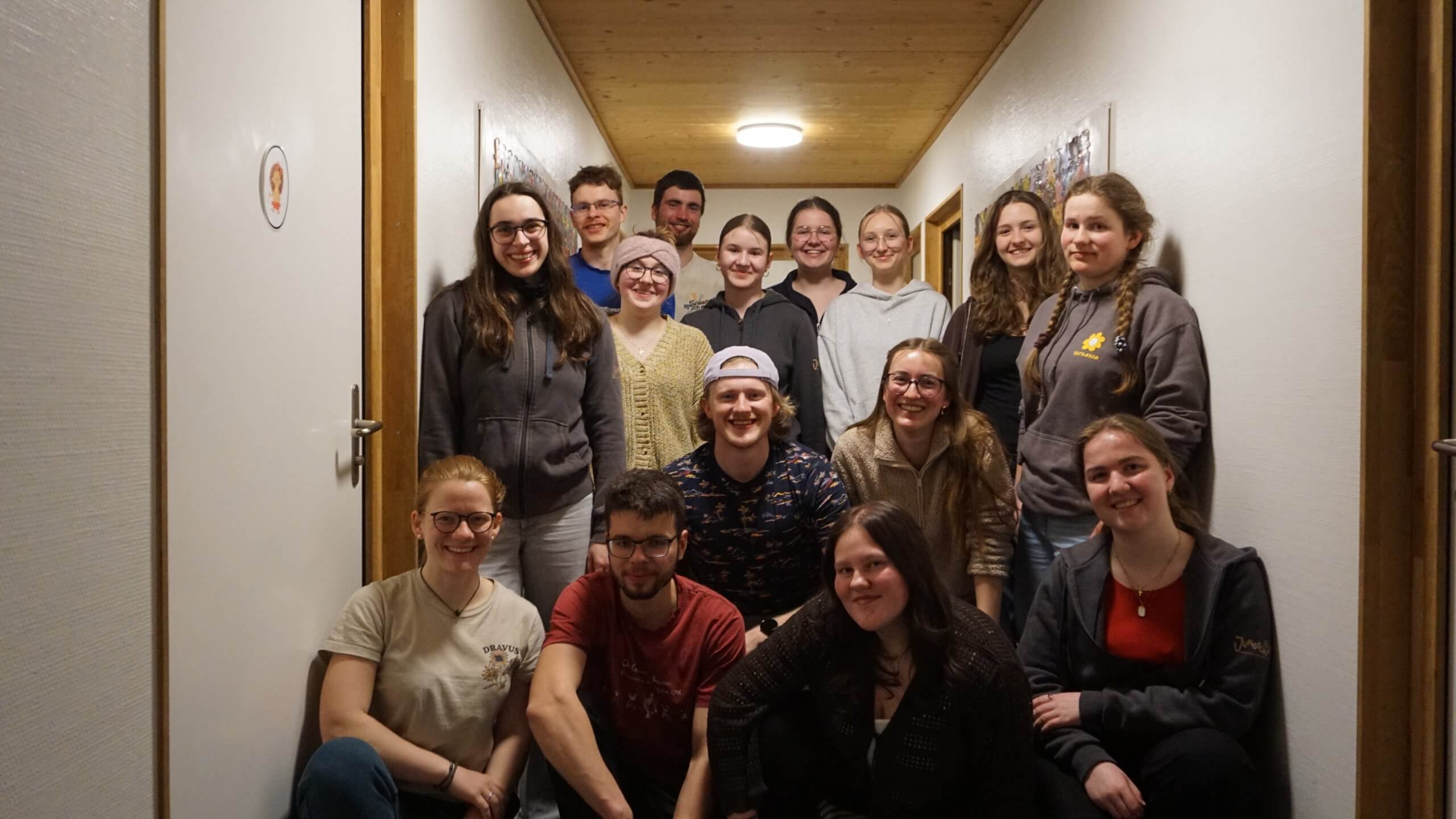 A large group of young adults posed together in a narrow hallway, smiling at the camera.